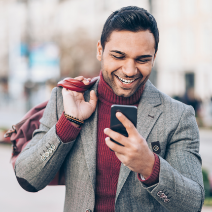 Man in a red shirt and bag smiles at his phone while walking down the street.